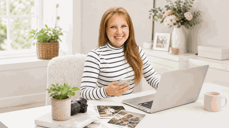 Miriam Meier sitzt lächelnd mit Smartphone und Laptop an einem hellen Schreibtisch.